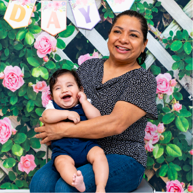 A woman holds a smiling baby in front of a floral backdrop.
