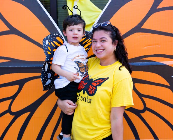 A woman in a yellow shirt holds a child in front of a monarch butterfly backdrop.